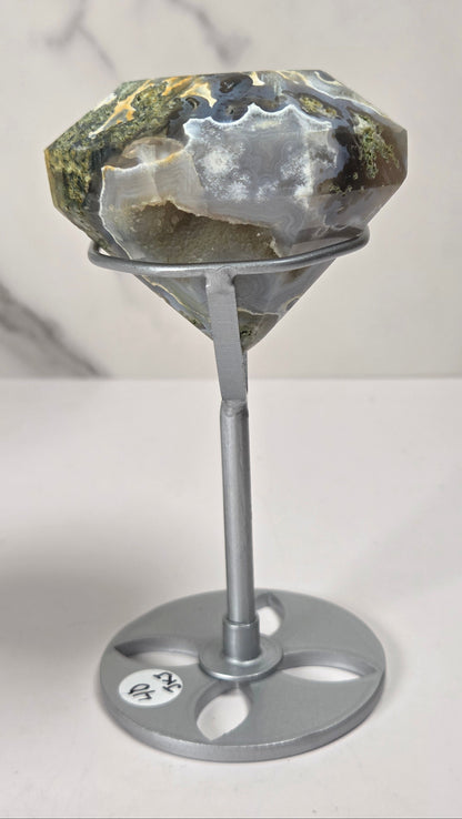 A large faceted gemstone with natural earthy tones of green, gray, and white, displayed on a silver metal stand against a light gray marble-patterned background.