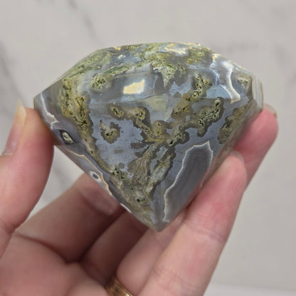 A hand holding a polished natural stone with a marbled pattern of gray, white, and olive-green, set against a light marble-patterned background.