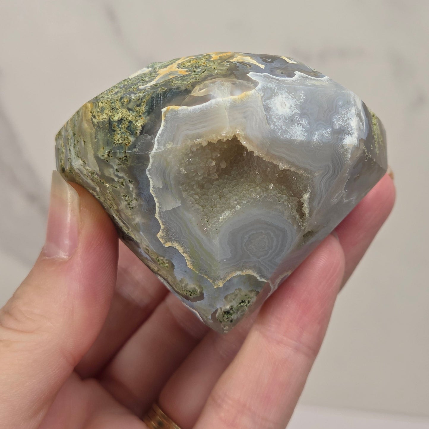 A hand holding a polished natural agate geode specimen with concentric band patterns in gray, white, olive green, and brown, against a light marble-patterned background.