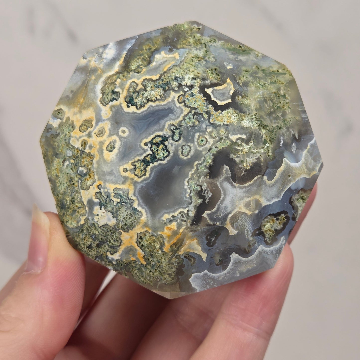 A hand holding a polished natural crystal or agate-like stone with swirling bands of gray, green, beige, and black, against a light marble-patterned background.