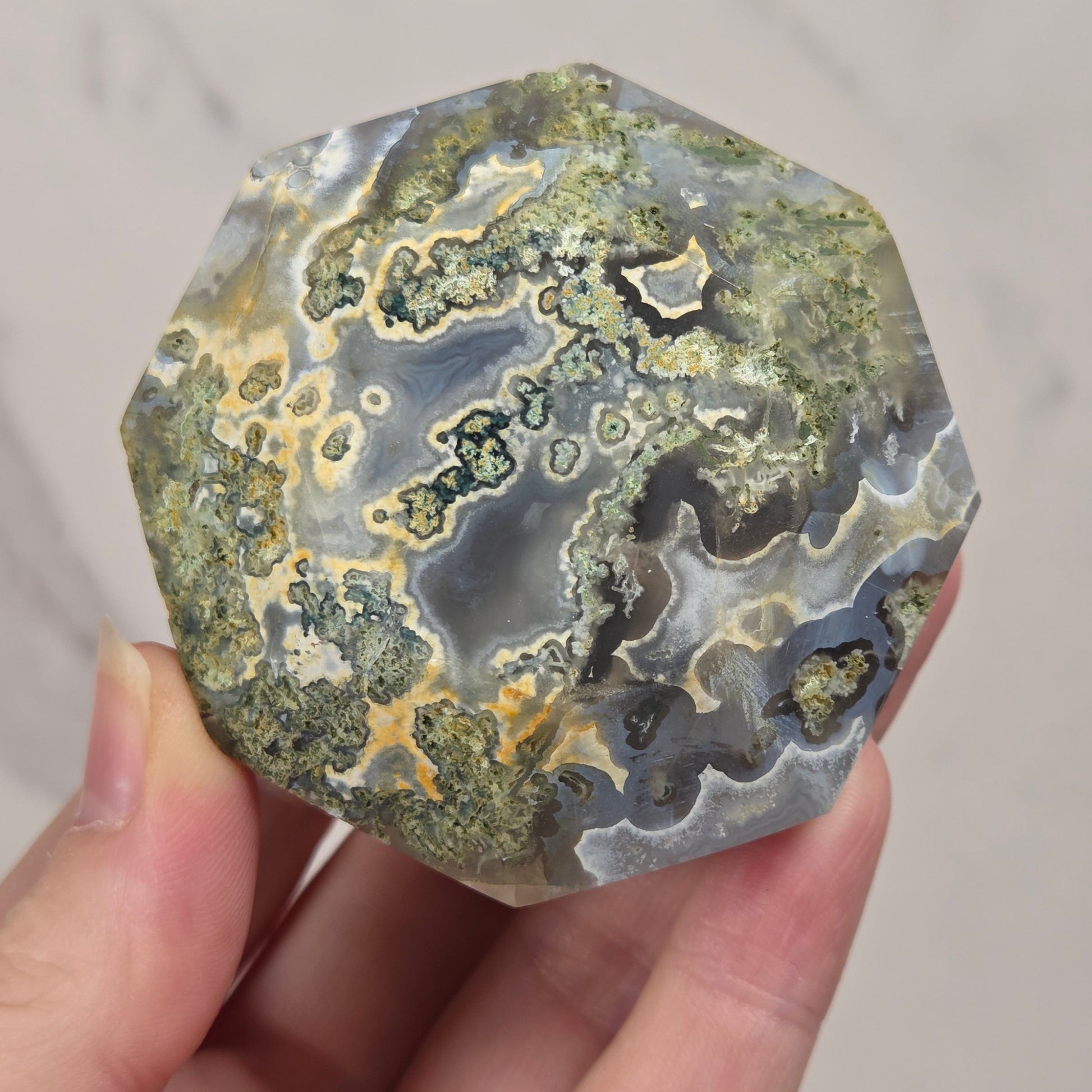 A hand holding a polished natural crystal or agate-like stone with swirling bands of gray, green, beige, and black, against a light marble-patterned background.