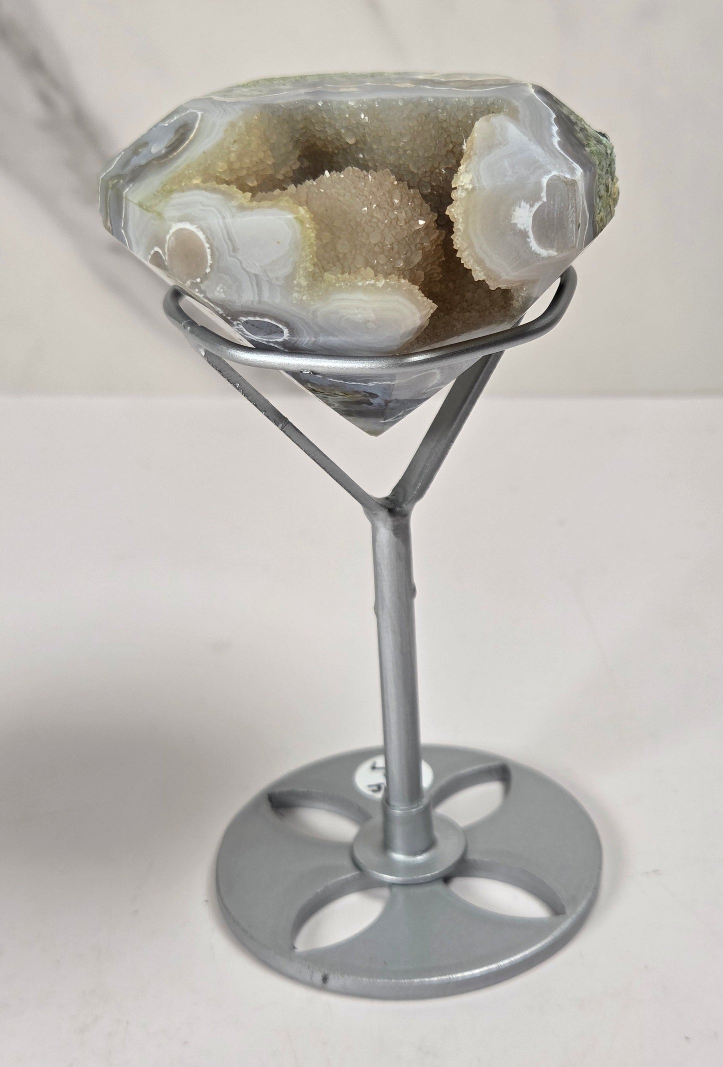 A faceted agate gemstone with natural banding in beige, gray, and white, displayed on a silver metal stand against a light marble-patterned background.