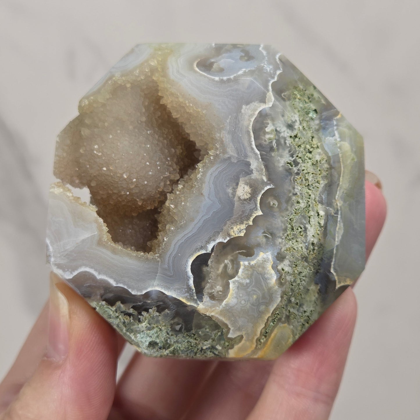 A hand holding a polished agate geode specimen with a natural cavity, showing layered bands of gray, white, beige, and greenish mineral deposits.
