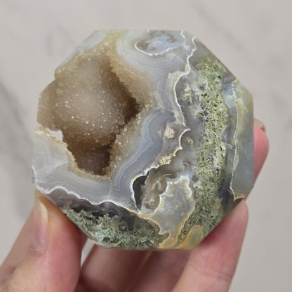 A hand holding a polished agate geode specimen with a natural cavity, showing layered bands of gray, white, beige, and greenish mineral deposits.