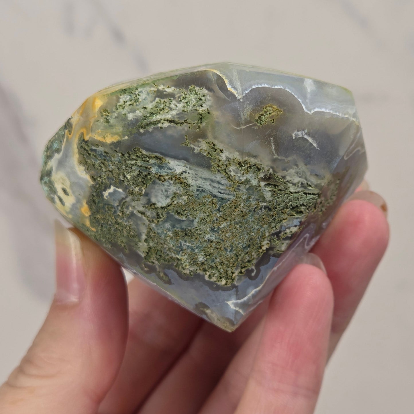 A hand holding a polished agate crystal cluster with green, gray, and gold banded patterns against a light marble background.