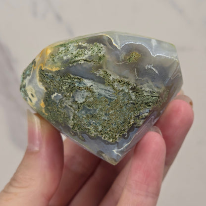 A hand holding a polished agate crystal cluster with green, gray, and gold banded patterns against a light marble background.