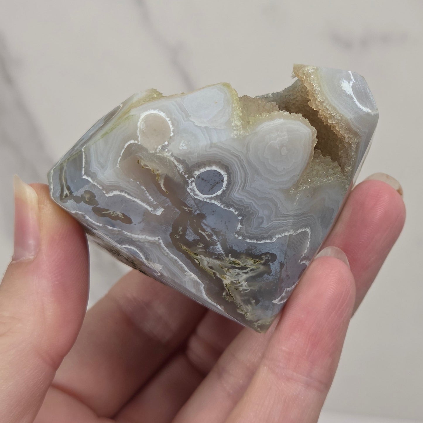 A hand holding a polished agate stone specimen with swirling bands of gray, white, beige, and brown, against a light marble-patterned background.