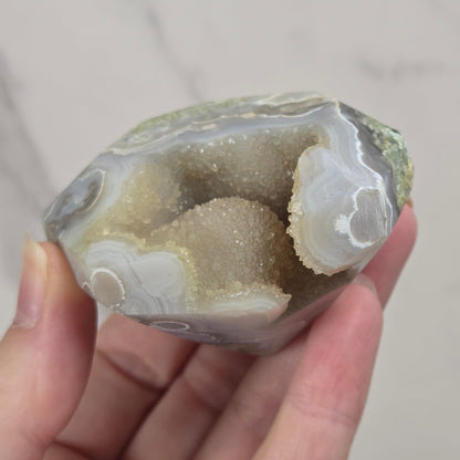 A hand holding a polished agate geode specimen with white and gray banded outer layers and a sandy beige inner cavity, set against a light marble-patterned background.