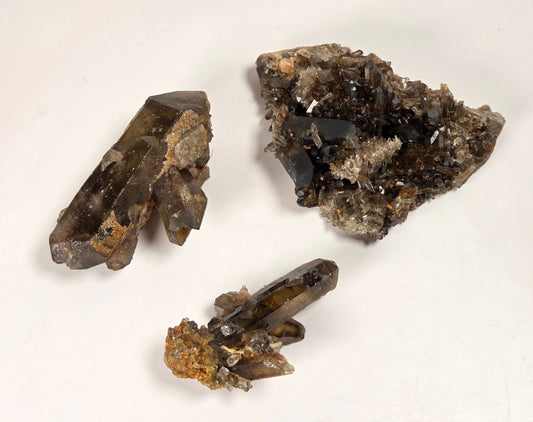Three mineral crystal clusters on a plain off-white background, including two translucent smoky brown crystal points and one rougher dark mineral specimen with lighter inclusions.