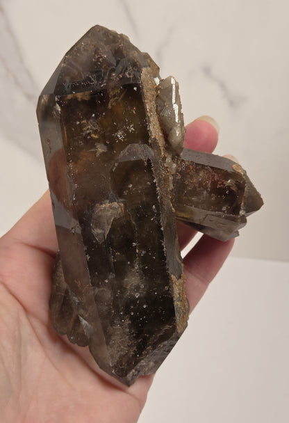 A person's hand holding a large natural smoky quartz crystal cluster with multiple points, showing its brown and gray translucent coloration against a light marble-patterned background.