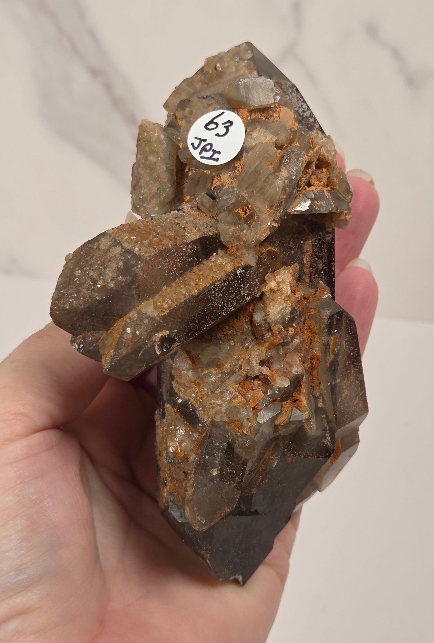 A hand holding a raw mineral specimen with dark gray to black crystals and orange-brown matrix, featuring a small white sticker with handwritten text reading '69 3oz'.