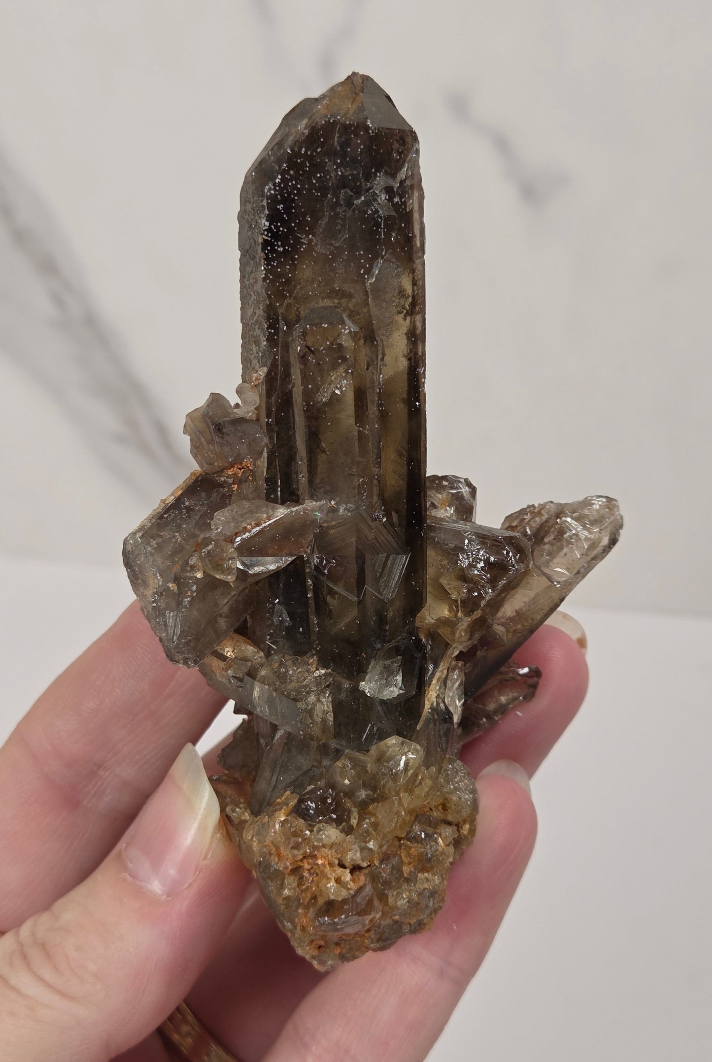 A hand holding a natural smoky quartz crystal cluster with a prominent central pointed crystal and a rough, earthy base, set against a light gray marble-patterned background.