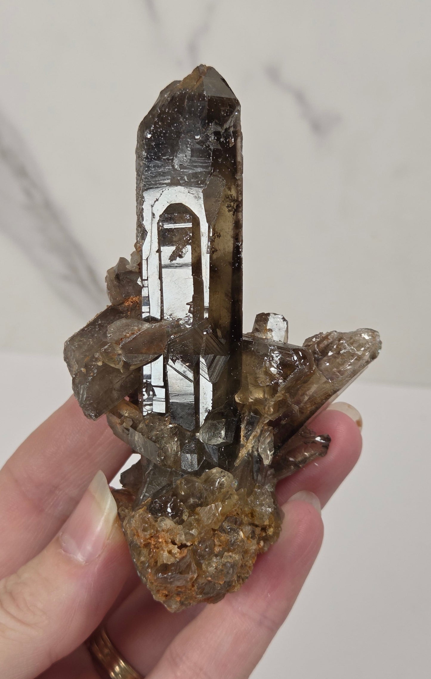 A hand holding a natural smoky quartz crystal cluster with a prominent pointed central tip, set against a light marble-patterned background.