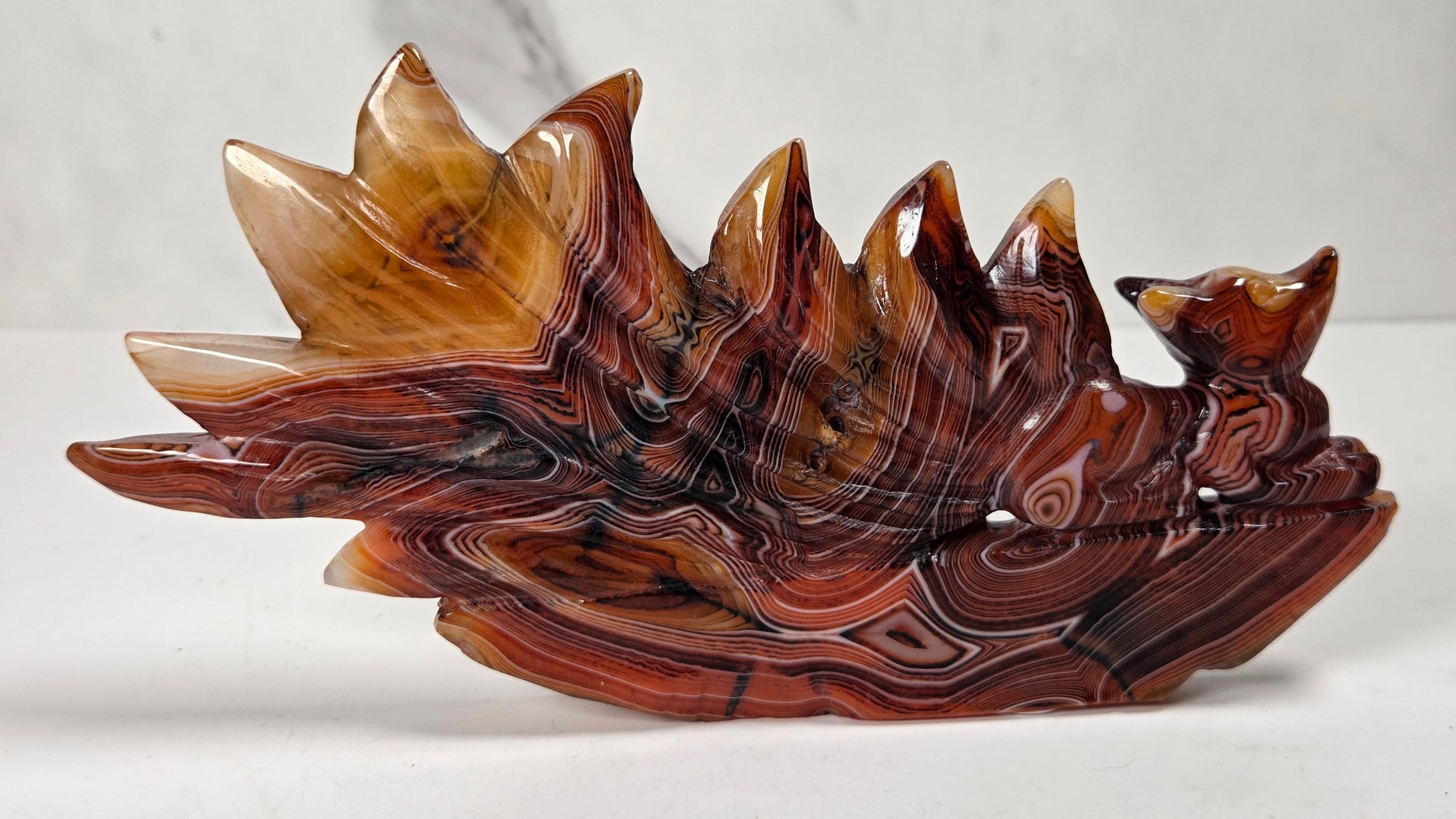 A carved decorative sculpture shaped like a fox riding a wave or mountain ridge, made from polished reddish-brown and amber stone with dark veining, photographed on a light surface against a pale marble-like background.