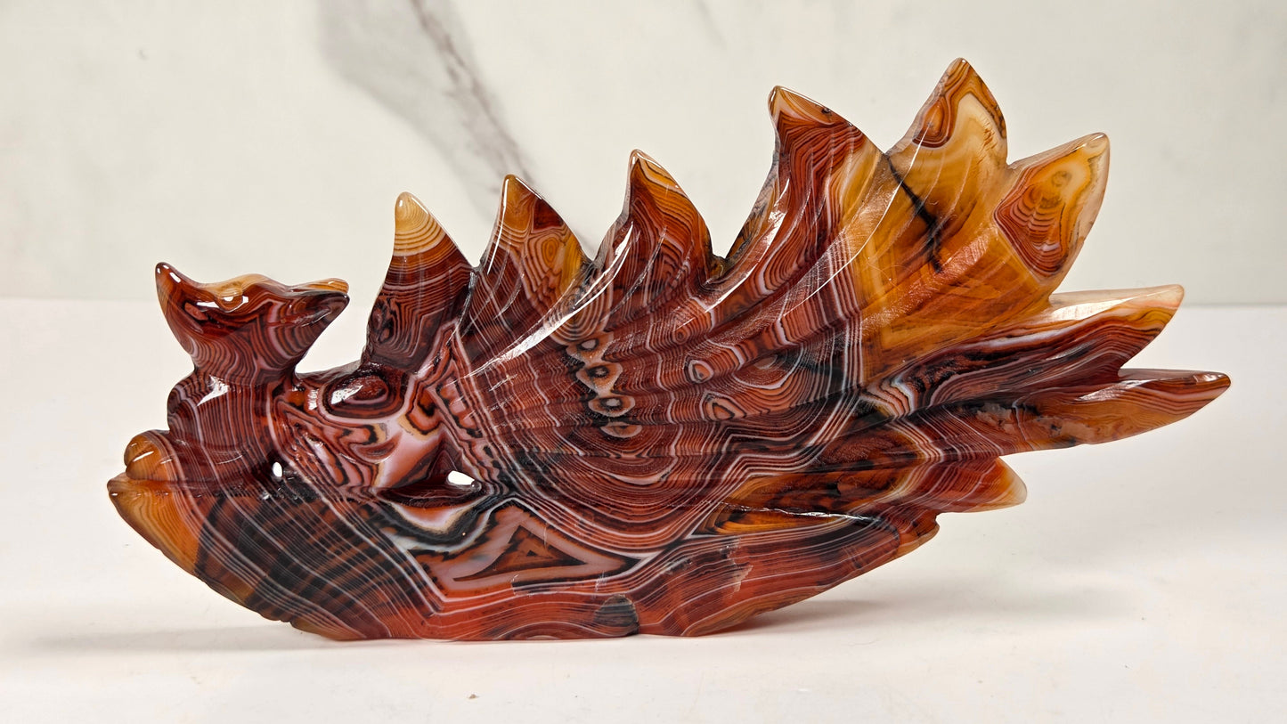A carved decorative sculpture resembling a stylized winged creature or dragon, made from polished red, orange, brown, and black marbled stone, displayed on a white surface against a light marble-patterned background.