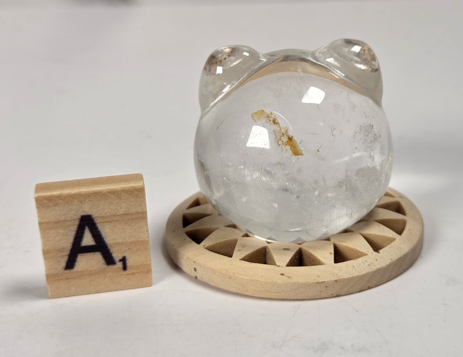A clear, rounded crystal or quartz bear figurine with small ears sits on a round wooden base, next to a small wooden letter tile showing the letter 'A'.