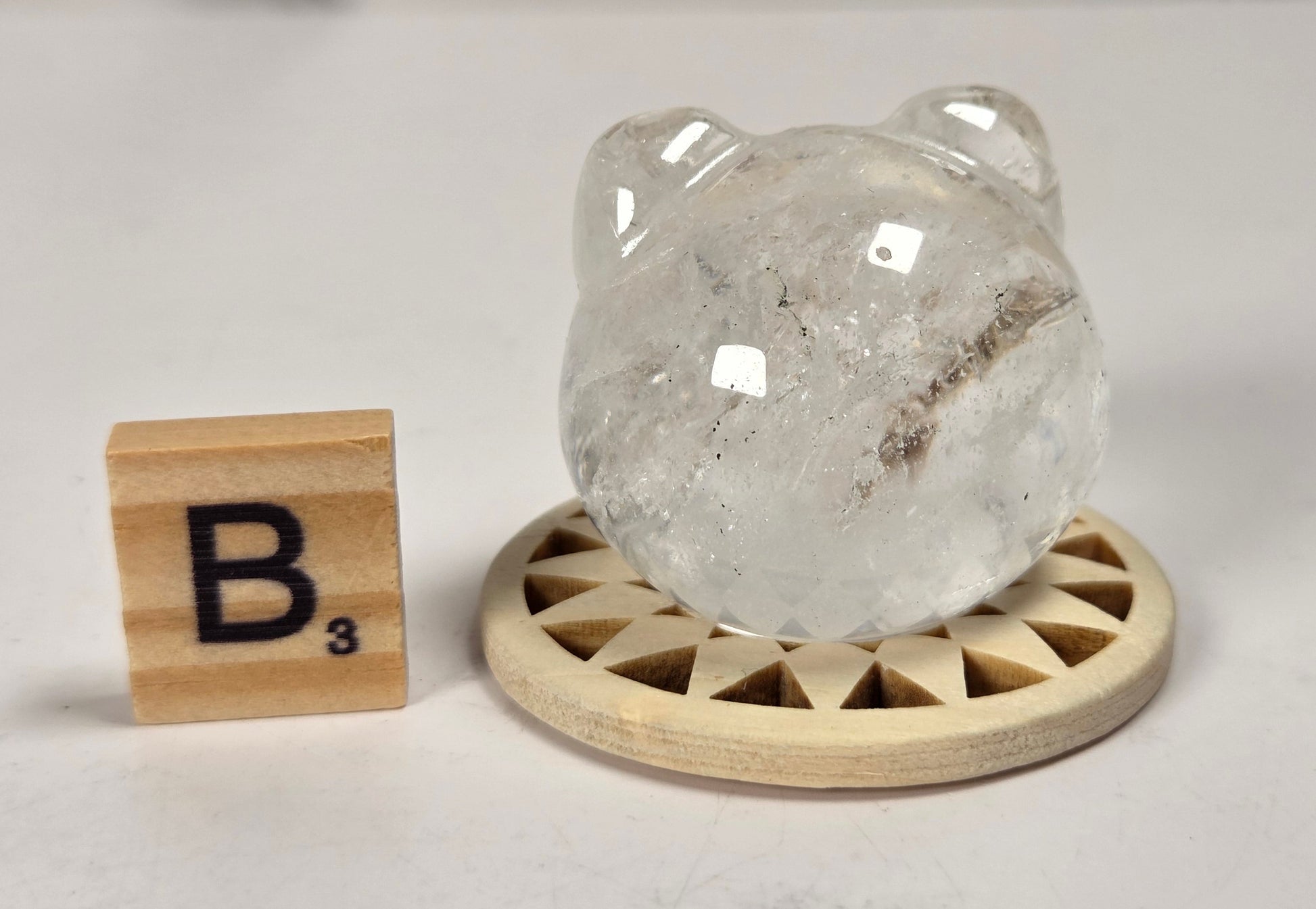 A clear, rounded crystal or stone sculpture with a bear-like shape, displayed on a small round wooden stand next to a wooden Scrabble tile showing the letter B.