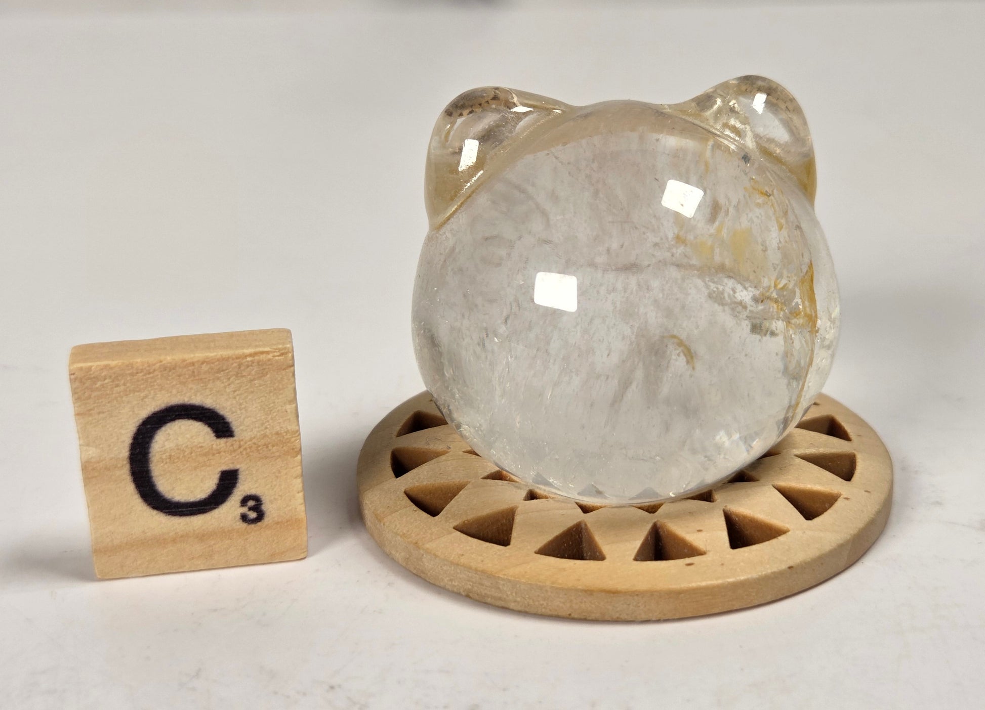 A clear quartz crystal sphere with natural inclusions, displayed on a round wooden stand next to a Scrabble tile marked C3, on a light marble surface.