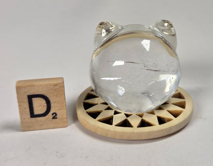 A clear crystal ball with a small frog head cutout at the top, displayed on a round wooden stand next to a wooden Scrabble tile marked D2, on a light gray surface.