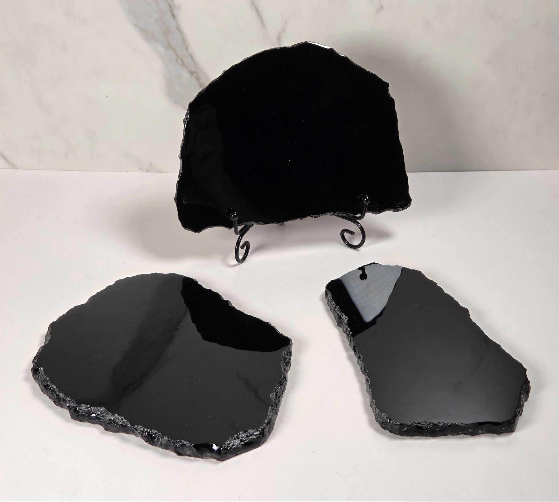 Three polished black stone slabs with rough natural edges displayed on a white surface, with one slab standing upright on a small black metal stand against a light marble-patterned background.