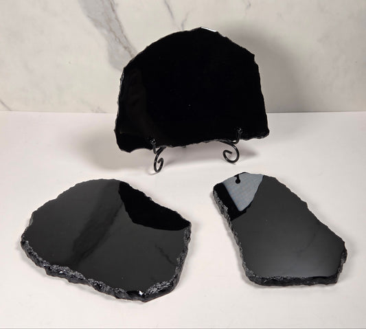 Three polished black stone slabs with rough natural edges displayed on a white surface, with one slab standing upright on a small black metal stand against a light marble-patterned background.