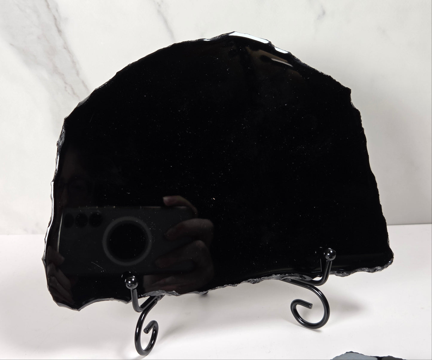 A large polished black stone slab with irregular edges, displayed on a black metal stand against a white marble-patterned background.