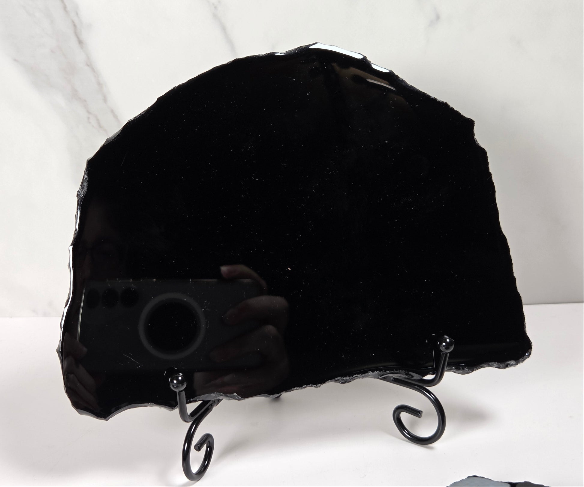 A large polished black stone slab with irregular edges, displayed on a black metal stand against a white marble-patterned background.