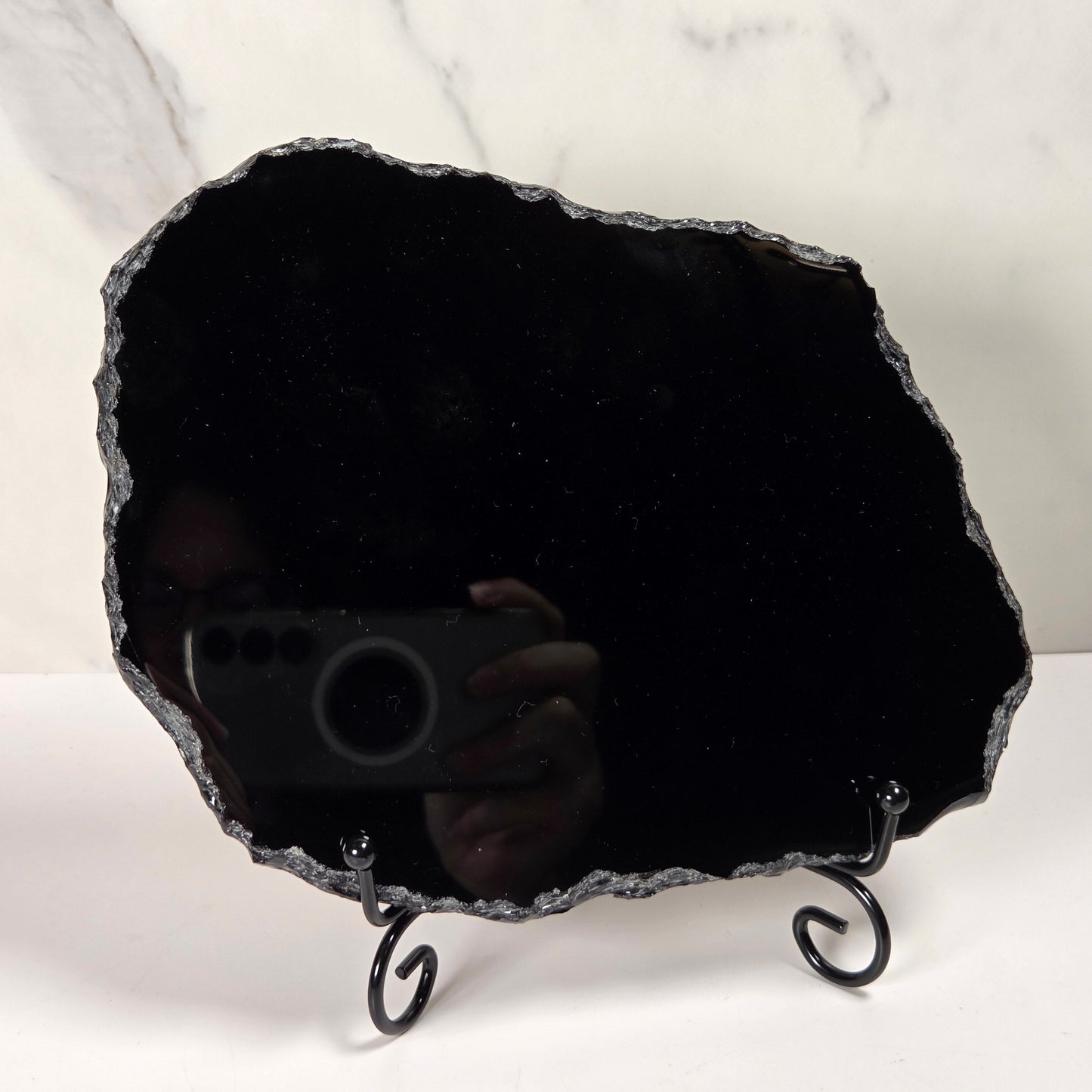 A raw black agate crystal cluster mirror with a black metal stand, photographed against a white marble-patterned background.