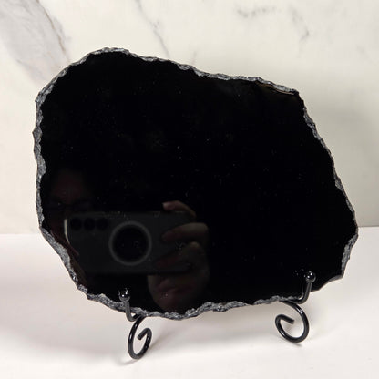 A raw black agate crystal cluster mirror with a black metal stand, photographed against a white marble-patterned background.