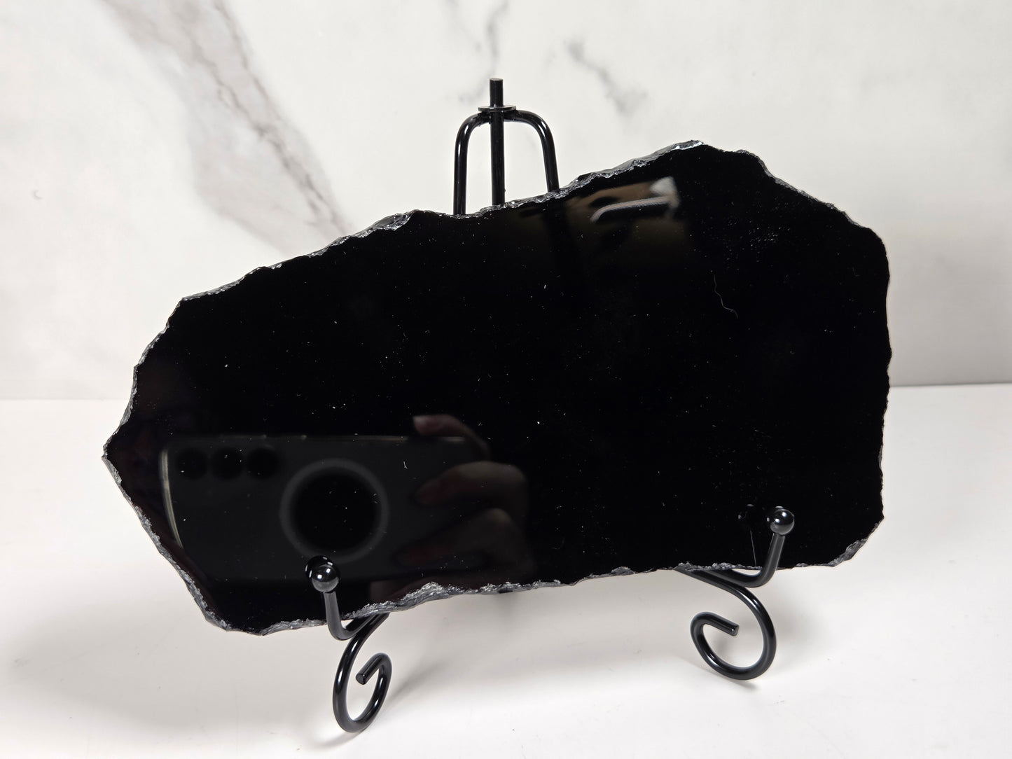 A black raw-edge mirror displayed on a black metal stand against a white marble-patterned background.
