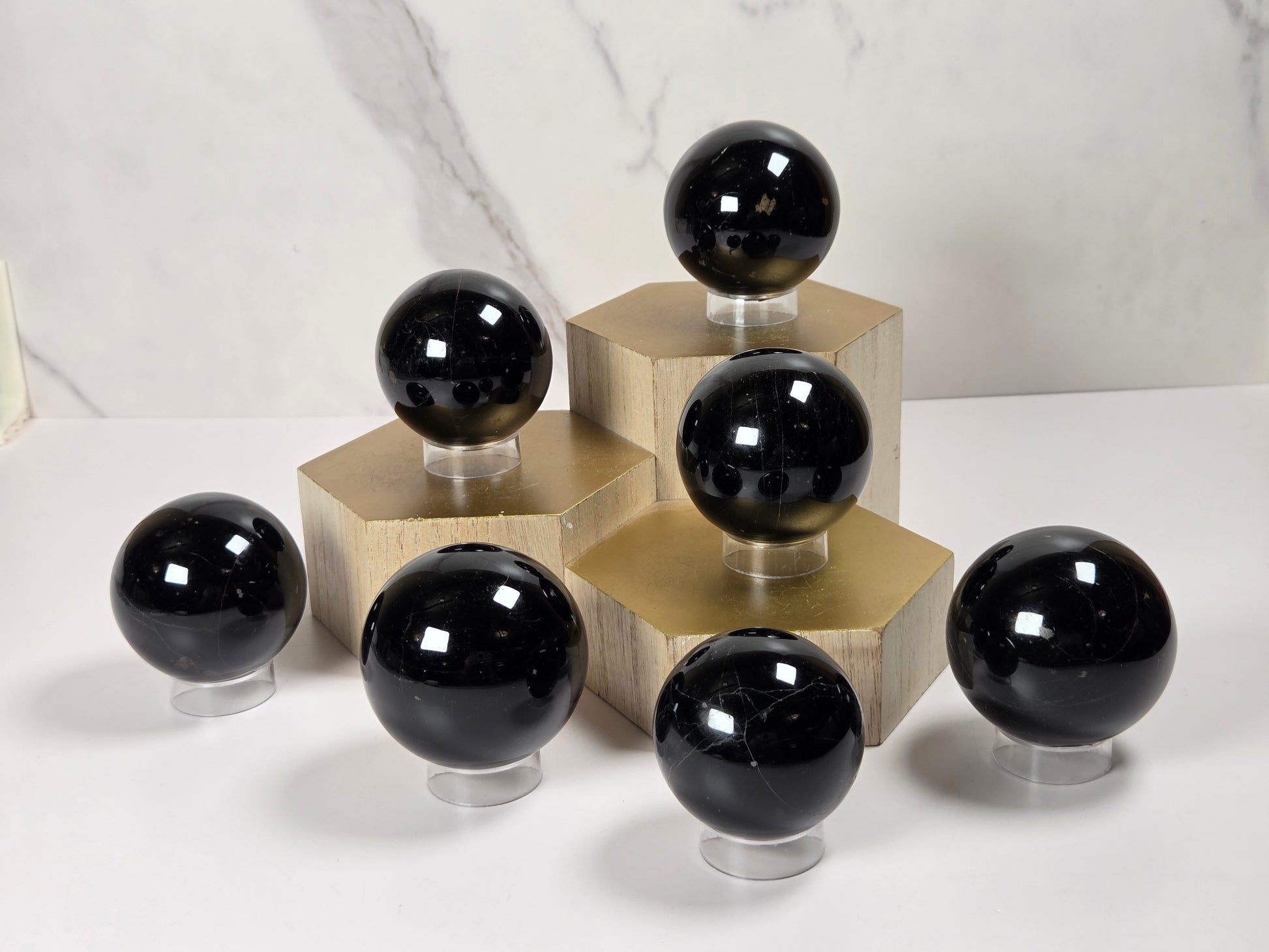 Seven polished black gemstone spheres of varying sizes displayed on clear acrylic stands, arranged on gold hexagonal pedestals against a white marble-patterned background.