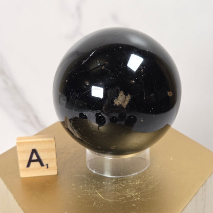 A polished black obsidian sphere with natural inclusions, displayed on a gold hexagonal pedestal next to a wooden letter tile showing the letter 'A'.