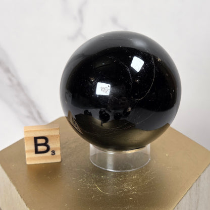 A glossy black polished stone sphere displayed on a clear stand atop a gold hexagonal pedestal, with a small wooden letter block showing 'B's' beside it against a light marble-pattern background.