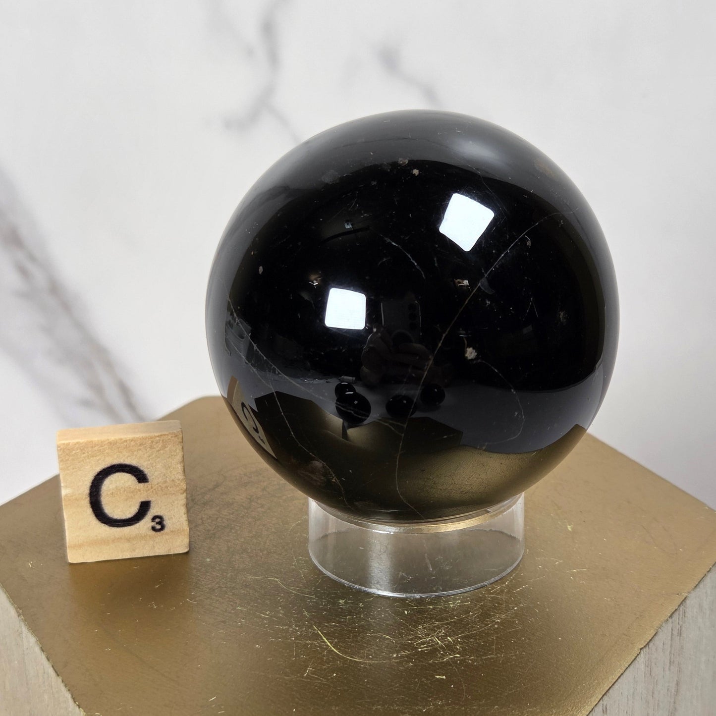 A glossy black crystal sphere displayed on a clear stand atop a gold hexagonal pedestal, with a small Scrabble tile showing 'C3' beside it against a light marble background.