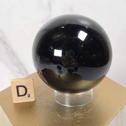 A glossy black polished stone sphere displayed on a clear acrylic stand atop a gold hexagonal pedestal, with a small wooden Scrabble tile showing the letter D² beside it against a light marble-look background.
