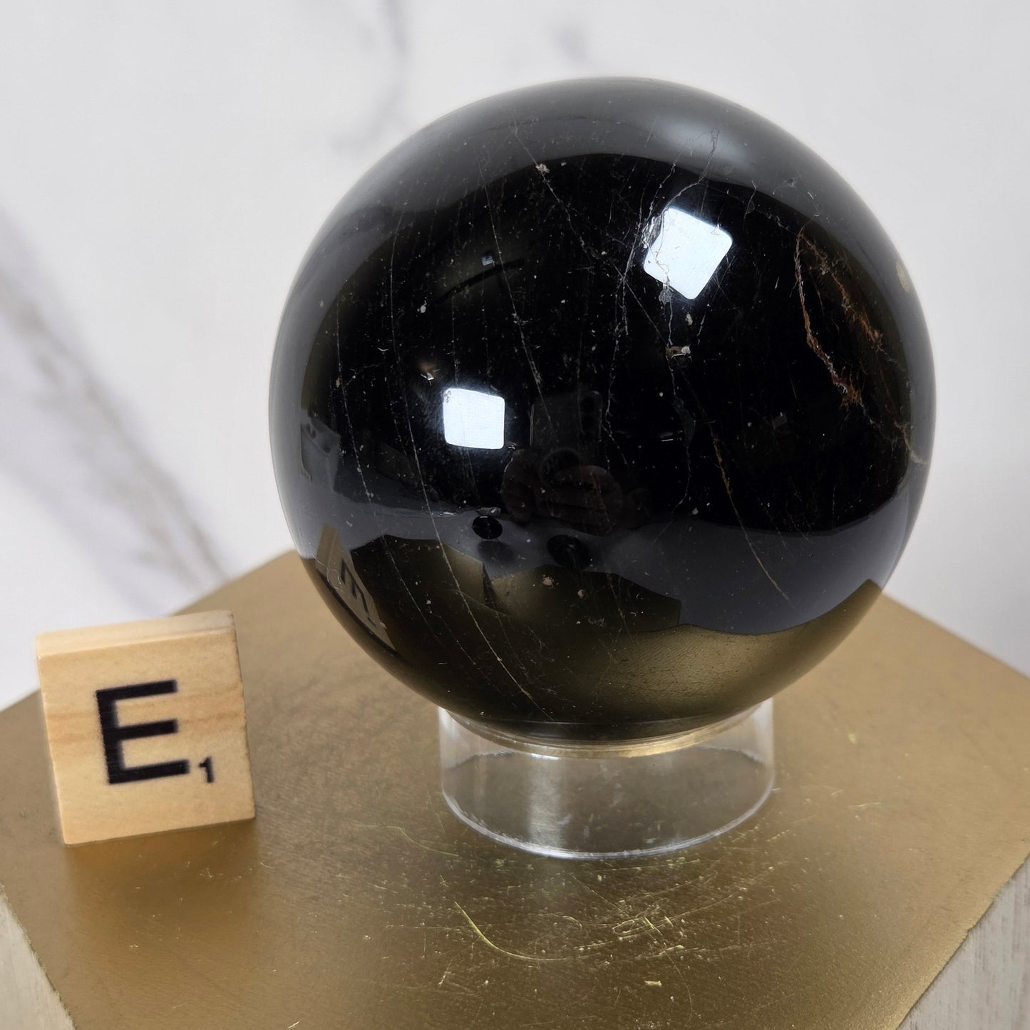 A glossy black polished crystal sphere displayed on a clear acrylic stand atop a gold hexagonal pedestal, with a small wooden block showing the letter 'E' beside it against a light marble-patterned background.