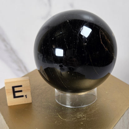 A glossy black polished crystal sphere displayed on a clear acrylic stand atop a gold hexagonal pedestal, with a small wooden block showing the letter 'E' beside it against a light marble-patterned background.