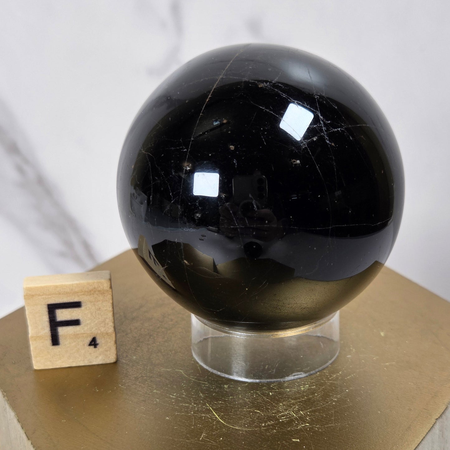 A glossy black polished stone sphere displayed on a clear acrylic stand, placed on a gold hexagonal pedestal next to a wooden letter block with the letter F.