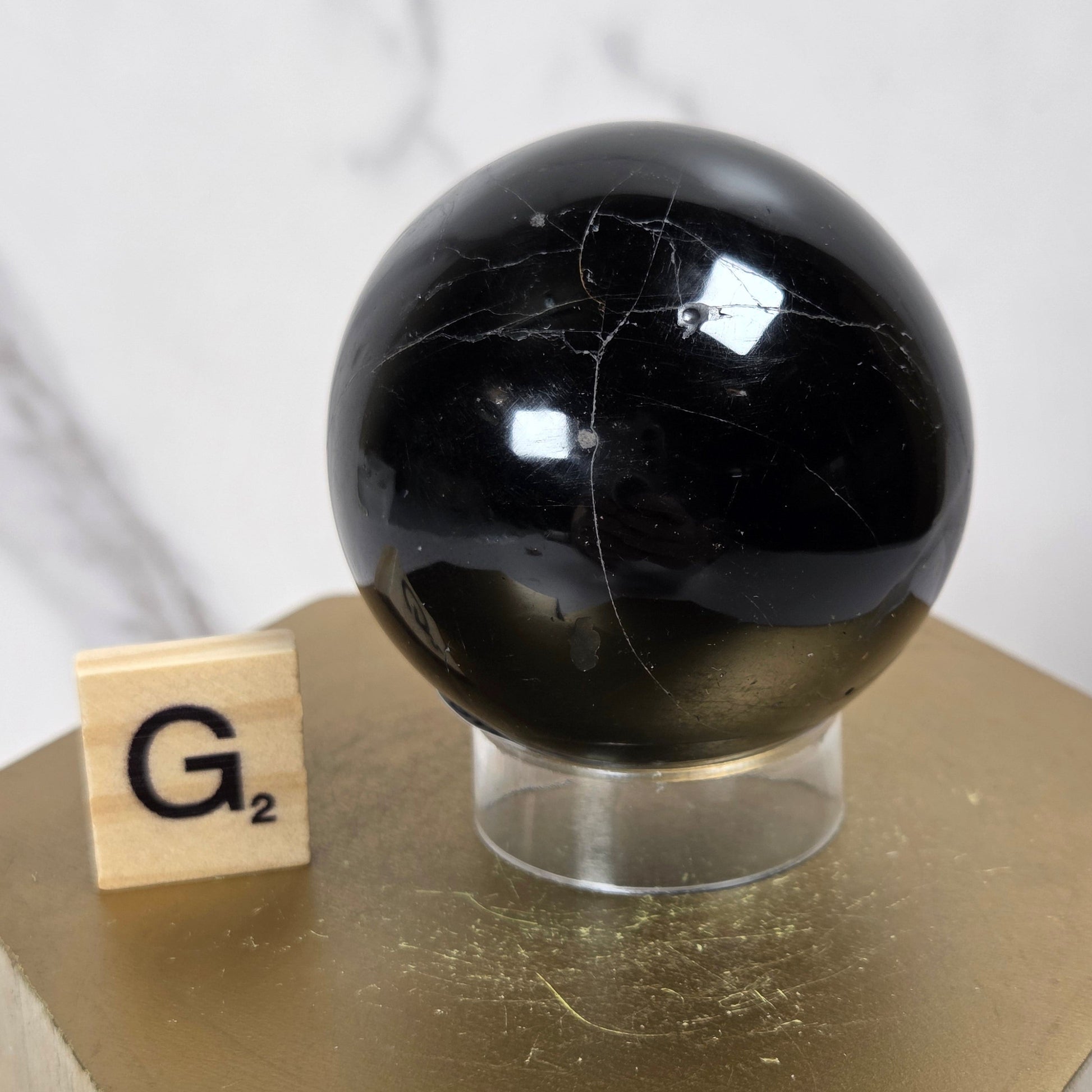 A polished black stone sphere with natural white veining, displayed on a clear acrylic stand next to a wooden Scrabble tile on a gold hexagonal pedestal.