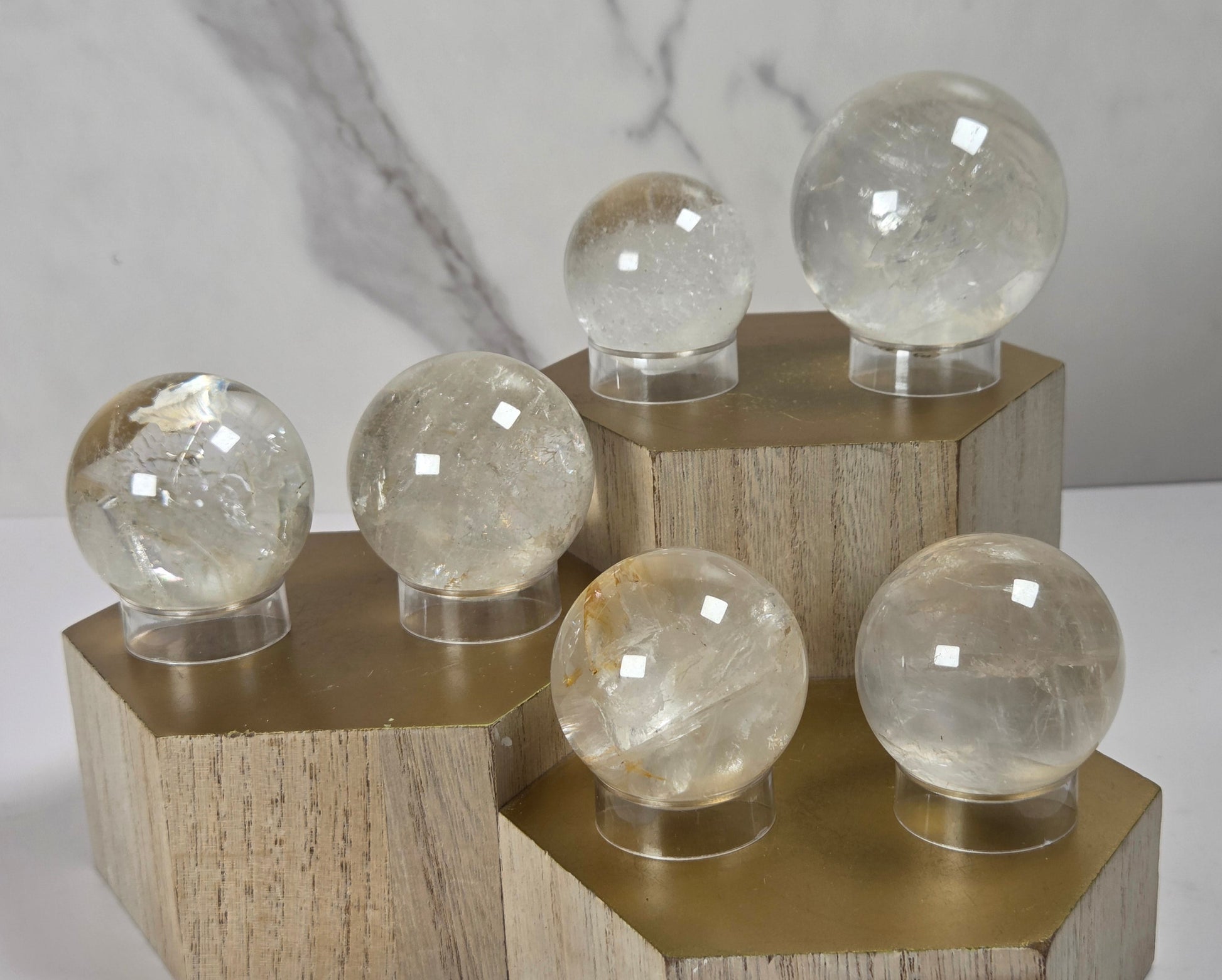 Set of clear quartz crystal spheres of varying sizes displayed on hexagonal gold-colored wooden pedestals against a light gray marble-patterned background.