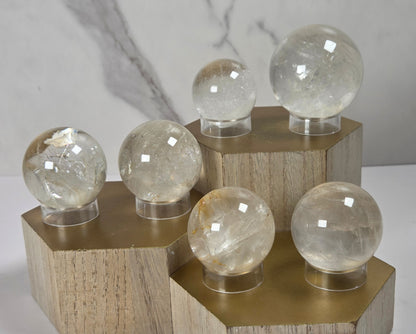 Set of clear quartz crystal spheres of varying sizes displayed on hexagonal gold-colored wooden pedestals against a light gray marble-patterned background.