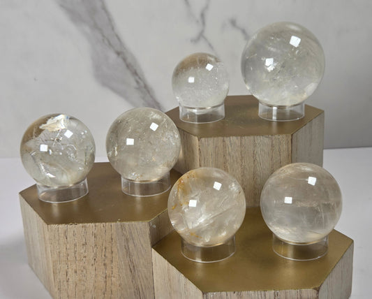 Set of clear quartz crystal spheres of varying sizes displayed on hexagonal gold-colored wooden pedestals against a light gray marble-patterned background.
