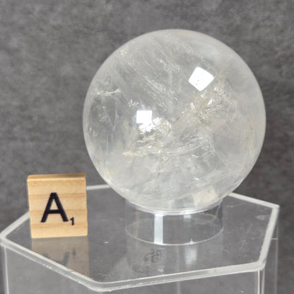 A large, clear quartz crystal sphere with visible internal inclusions, displayed on a clear acrylic hexagonal stand next to a wooden Scrabble tile showing the letter 'A'. The background is a textured gray wall.
