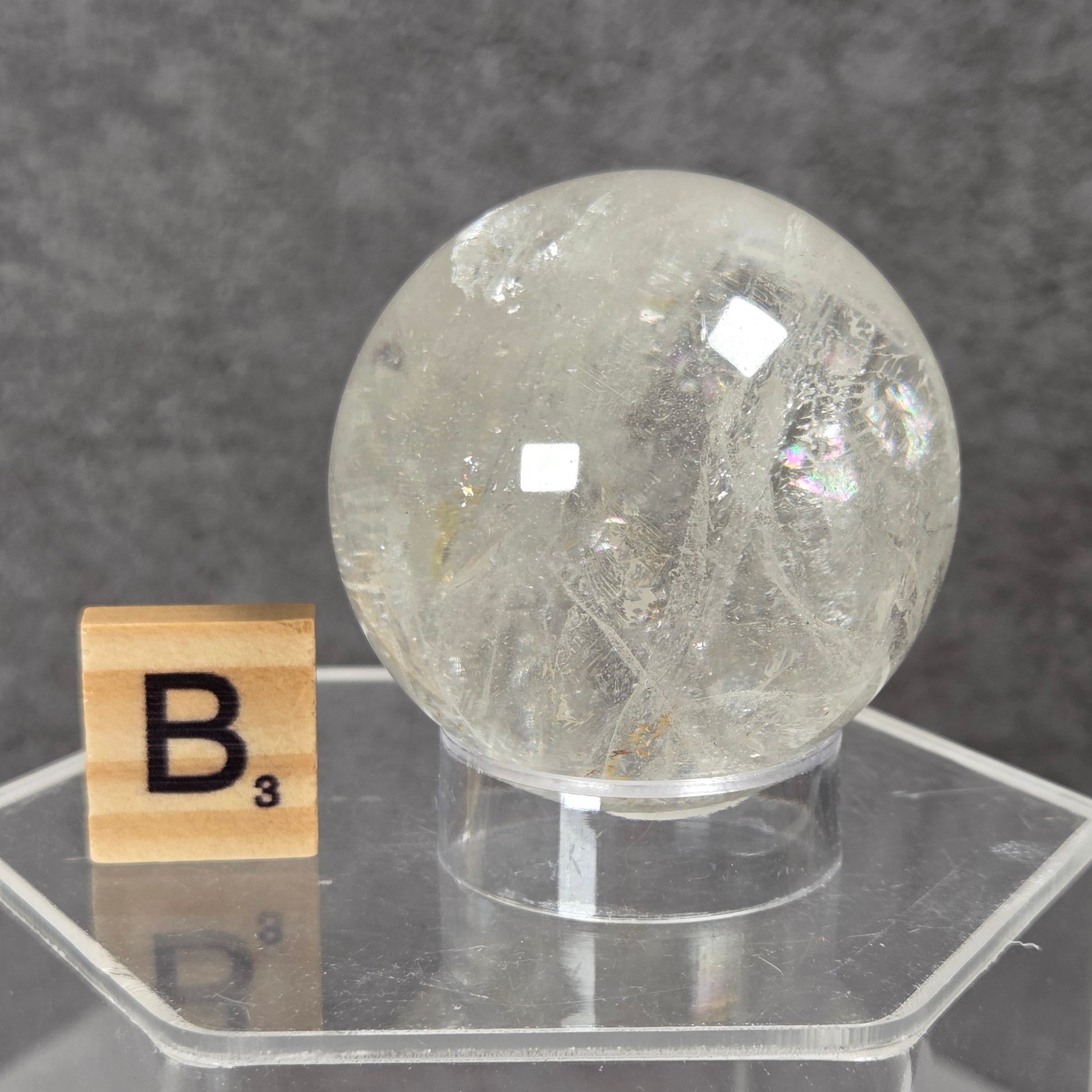 A clear faceted crystal sphere displayed on a transparent acrylic stand, with a small Scrabble tile showing the letter B next to it, against a dark gray textured background.