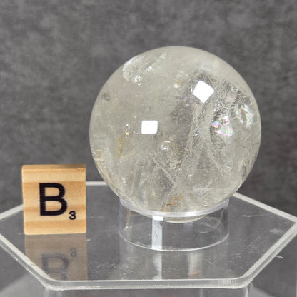A clear faceted crystal sphere displayed on a transparent acrylic stand, with a small Scrabble tile showing the letter B next to it, against a dark gray textured background.