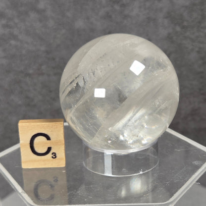 A clear quartz crystal sphere on a transparent acrylic display stand, with a small Scrabble tile showing the letter C next to it, set against a dark gray textured background.