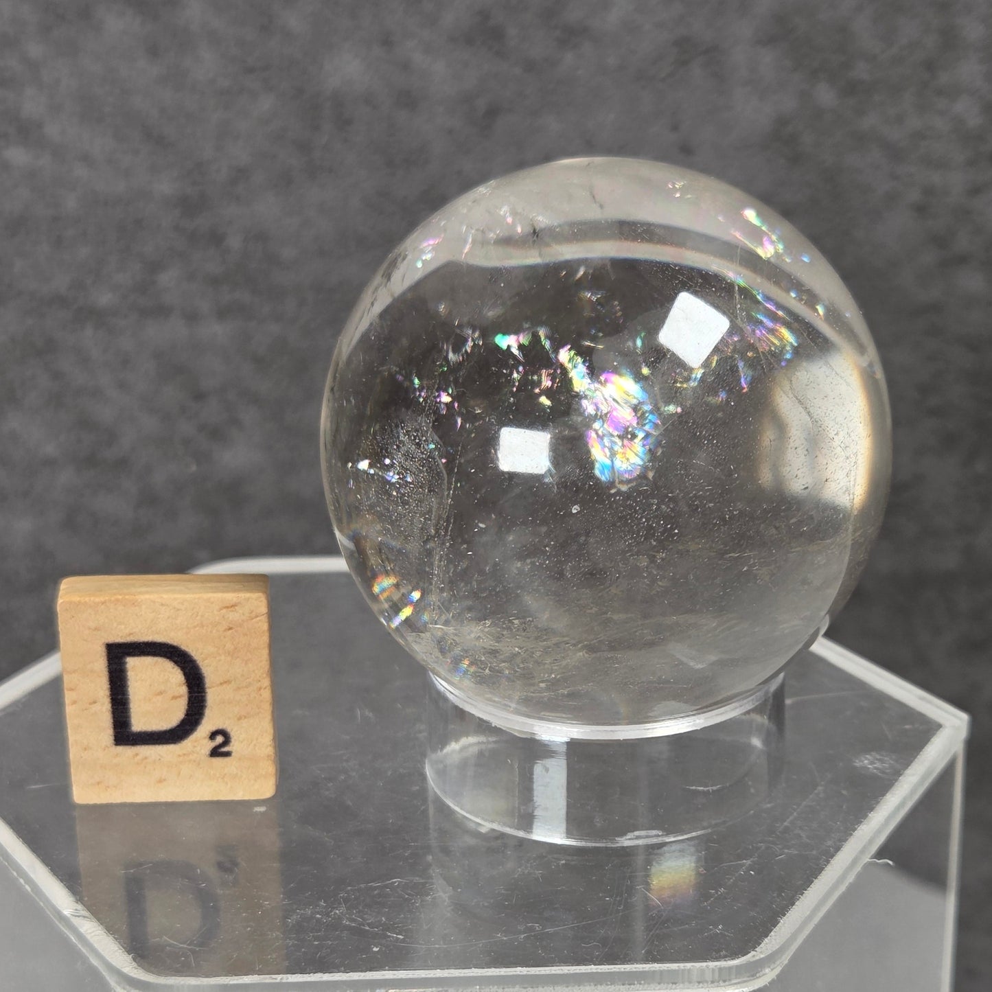 A clear glass or acrylic decorative sphere displayed on a transparent acrylic stand, with a small Scrabble tile showing D² beside it against a dark gray textured background.