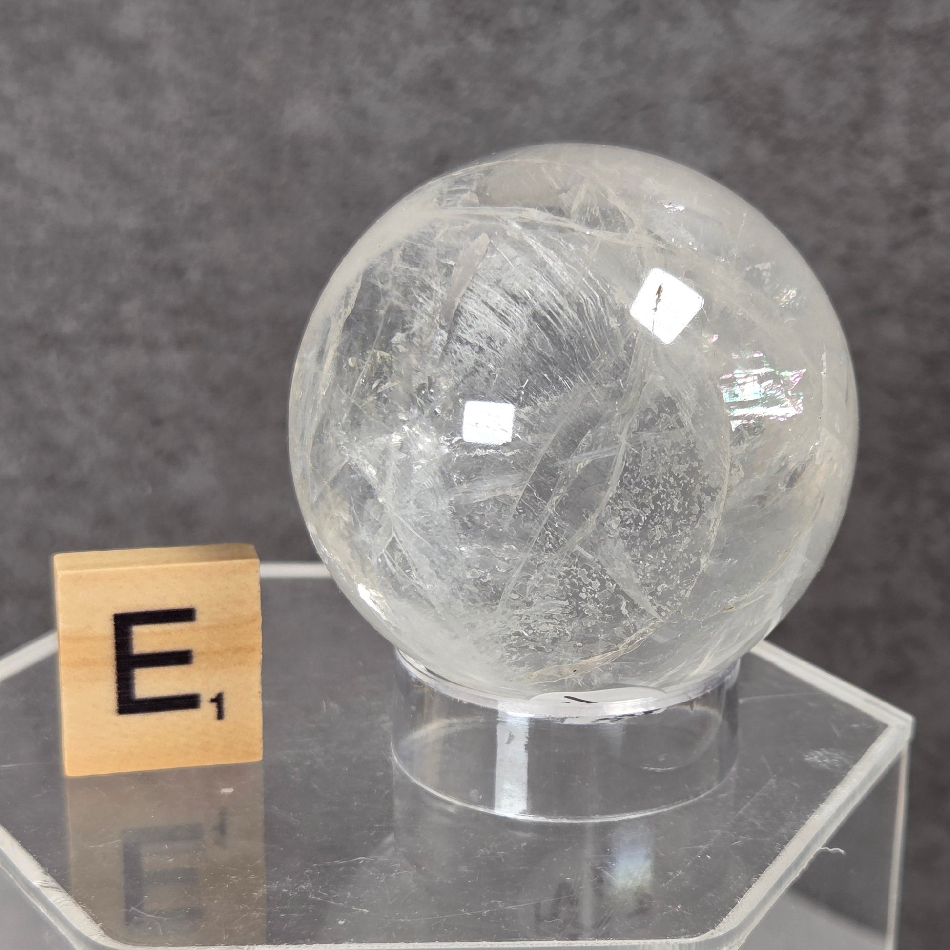 A clear, spherical crystal formation with internal striations sits on a transparent acrylic display base next to a small wooden Scrabble tile showing the letter E, set against a dark gray textured background.