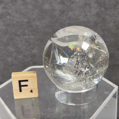 A clear, transparent spherical crystal or glass orb with internal inclusions, displayed on a clear acrylic hexagonal pedestal next to a wooden Scrabble tile showing the letter F.