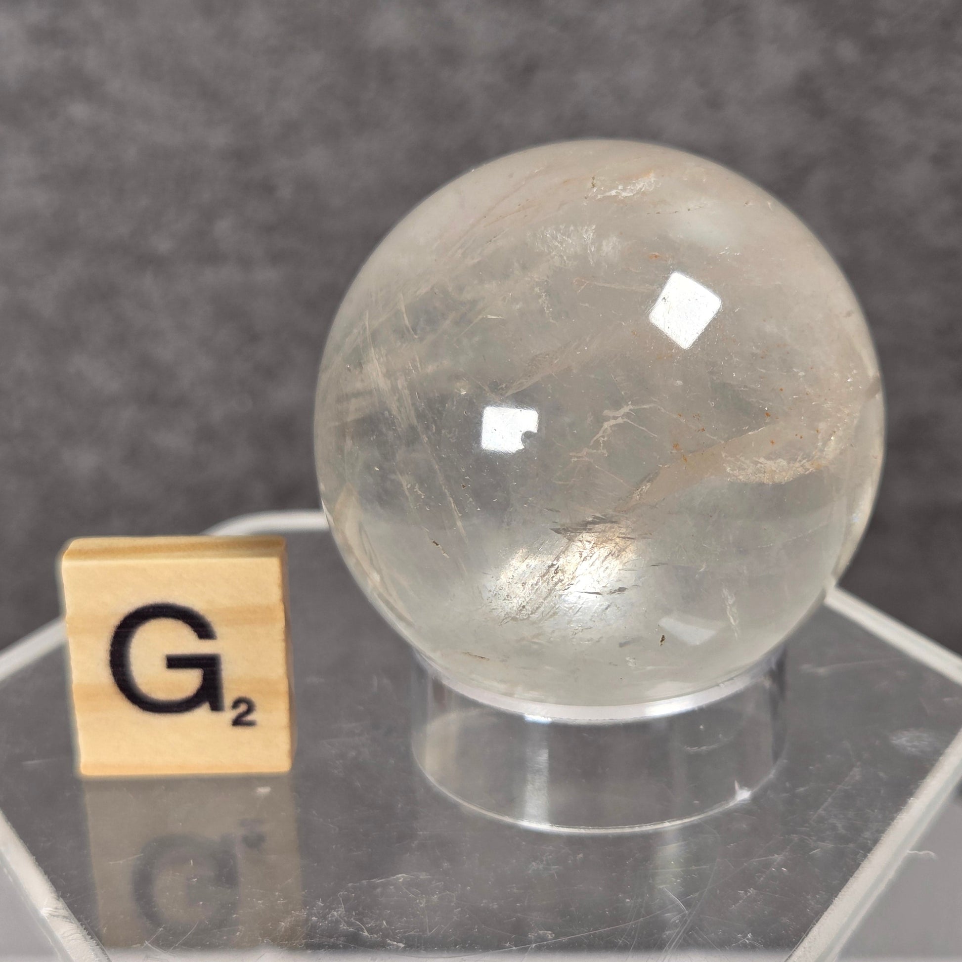 A clear, rounded crystal sphere with internal inclusions, displayed on a transparent acrylic hexagonal stand next to a wooden Scrabble tile showing the letter G2, set against a dark gray textured background.